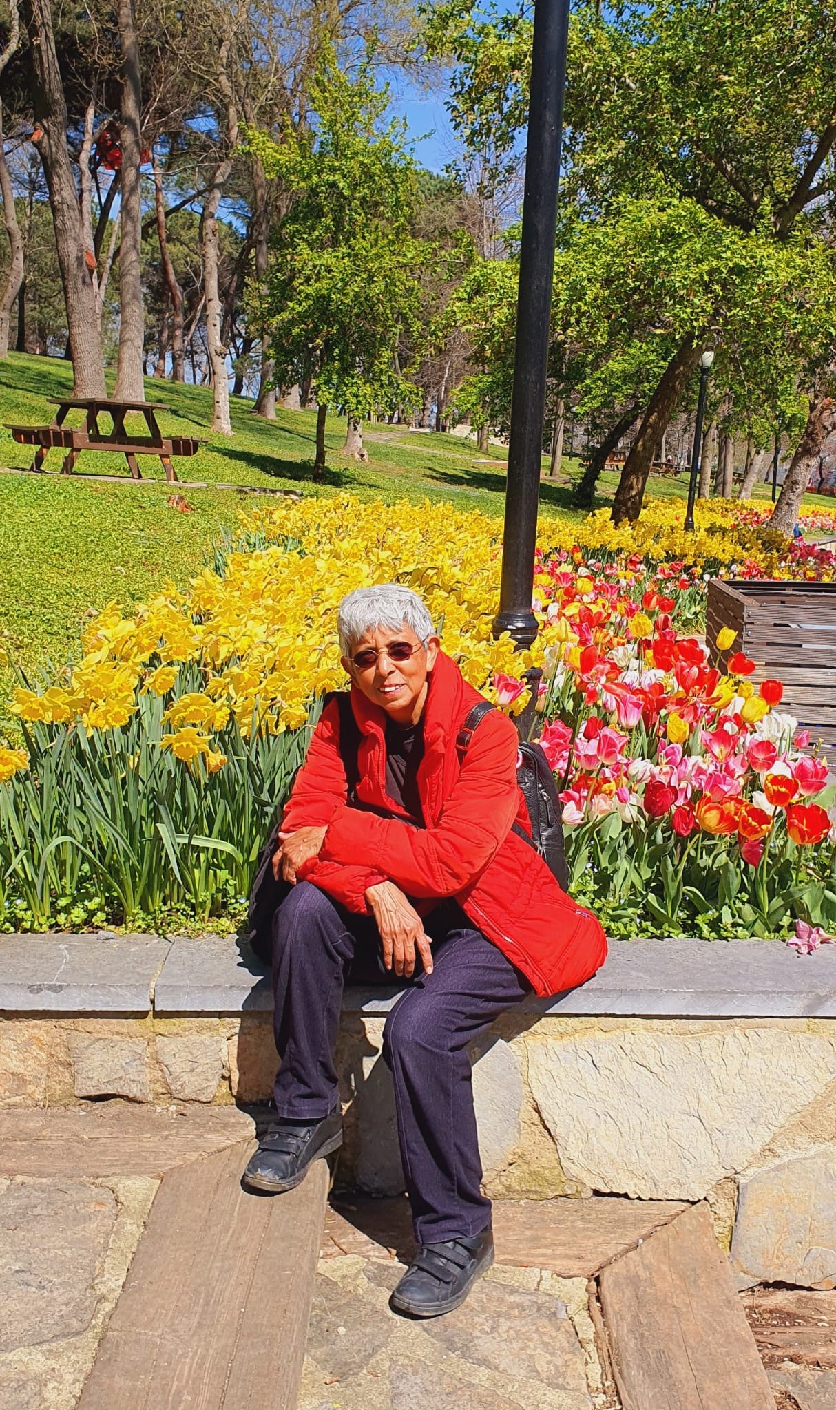 Smiling woman posing in a seated position, with a park behind her and a long bed of red and yellow tulips.